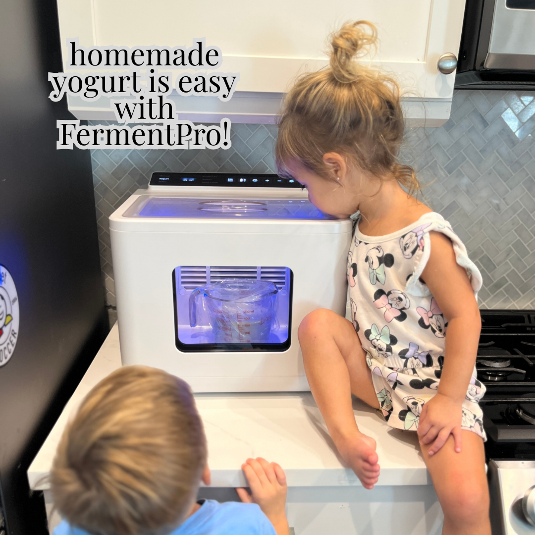 Two children looking into a white appliance with a blue light, likely a yogurt maker, in a kitchen setting.